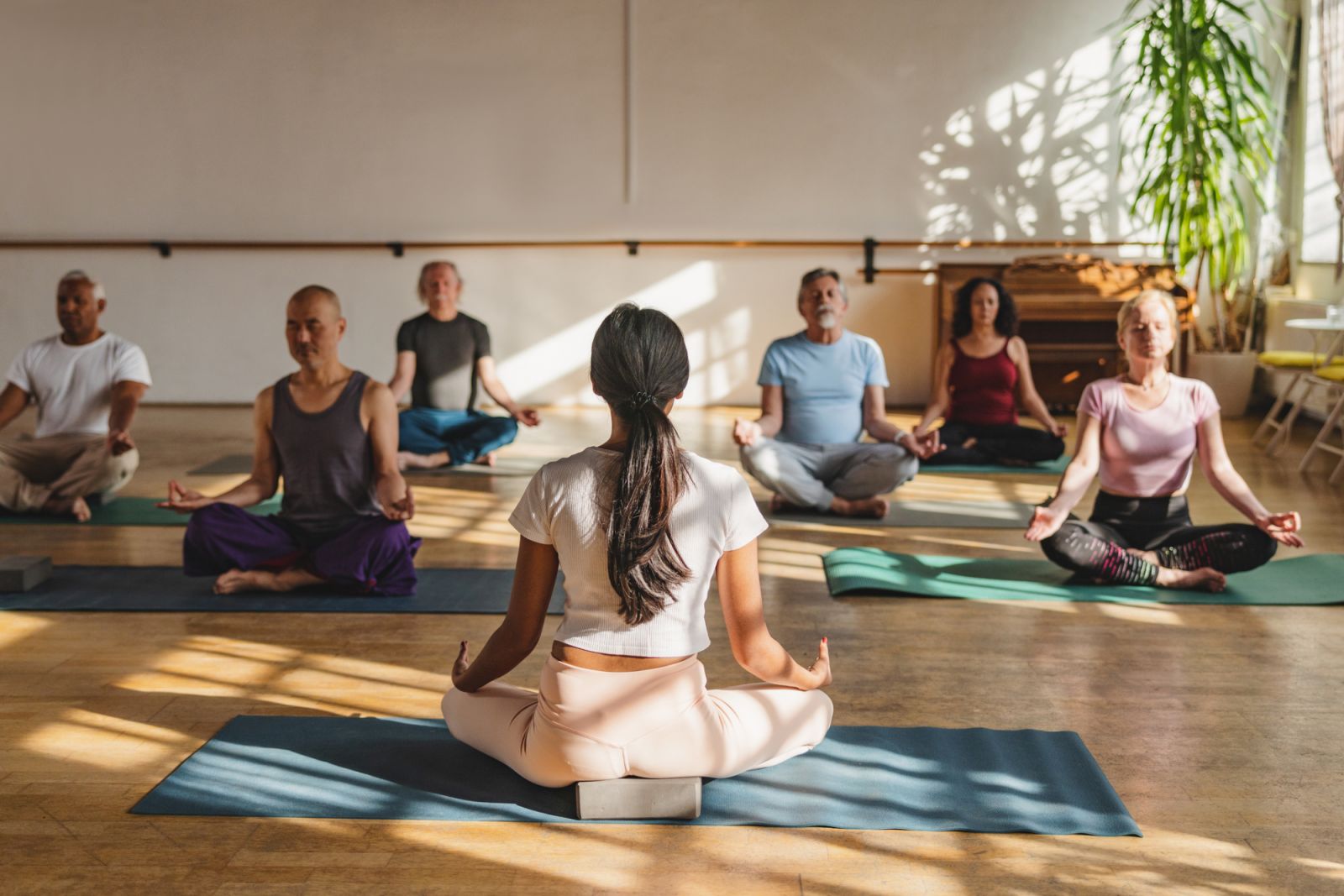 Full length front shot with blurred background of multicultural mature yoga participants seated on their mats meditating while following her female instructor indoors during a sunny day.