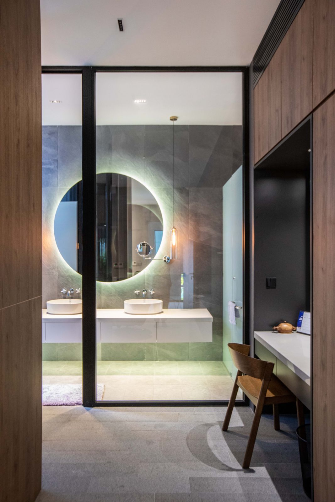 The bathroom's minimalist design features a striking backlit circular mirror, twin vessel sinks, and floor-to-ceiling glass partitions, demonstrating Formzero's attention to detail and harmonious material selection of wood, glass, and stone."