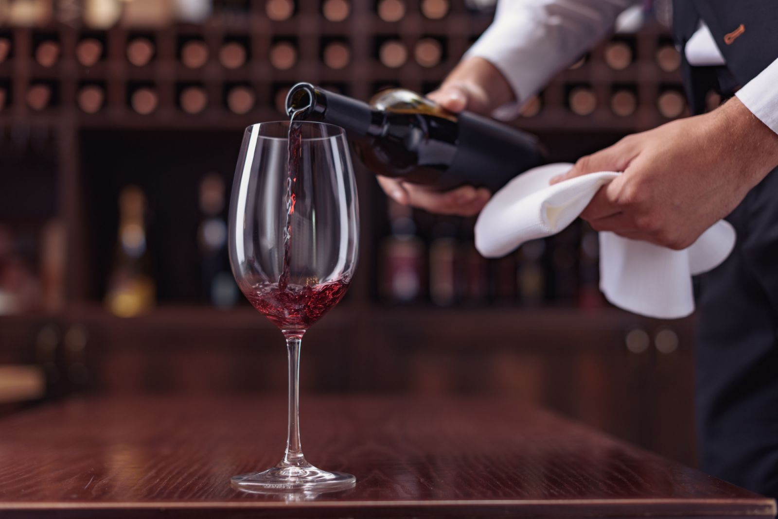 Cropped view sommelier pouring red wine from bottle into glass at table in cellar