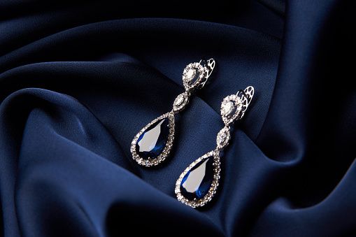 Two Golden sapphire earrings with small diamonds. Pair of platinum earring with sapphire gemstone on blue satin background. Luxury female jewelry, close-up
