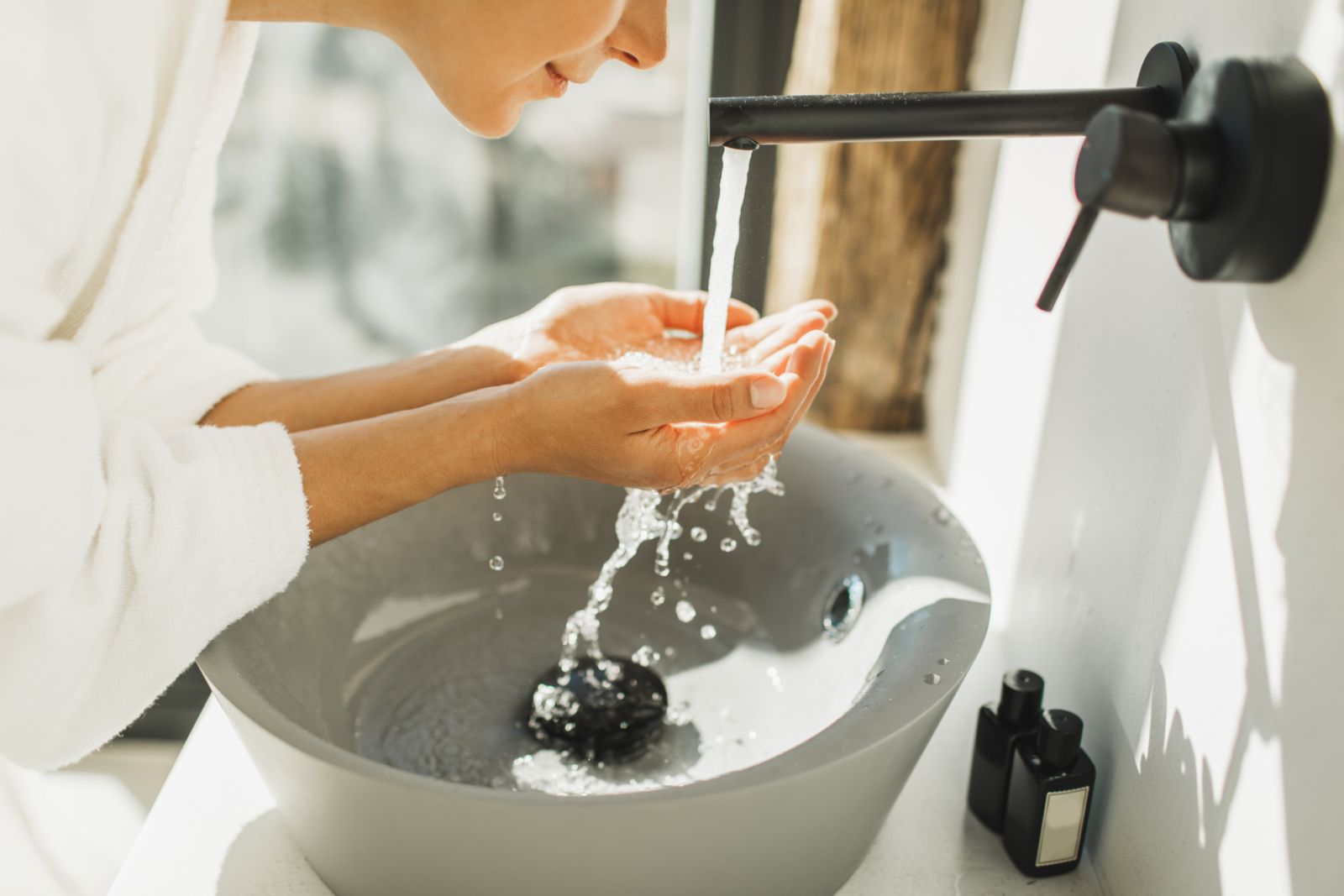 Spa treatment. Bathroom with white walls full of sun light. Black metal furniture. Grey stone sink. Person in bathrobe. Trendy interior design