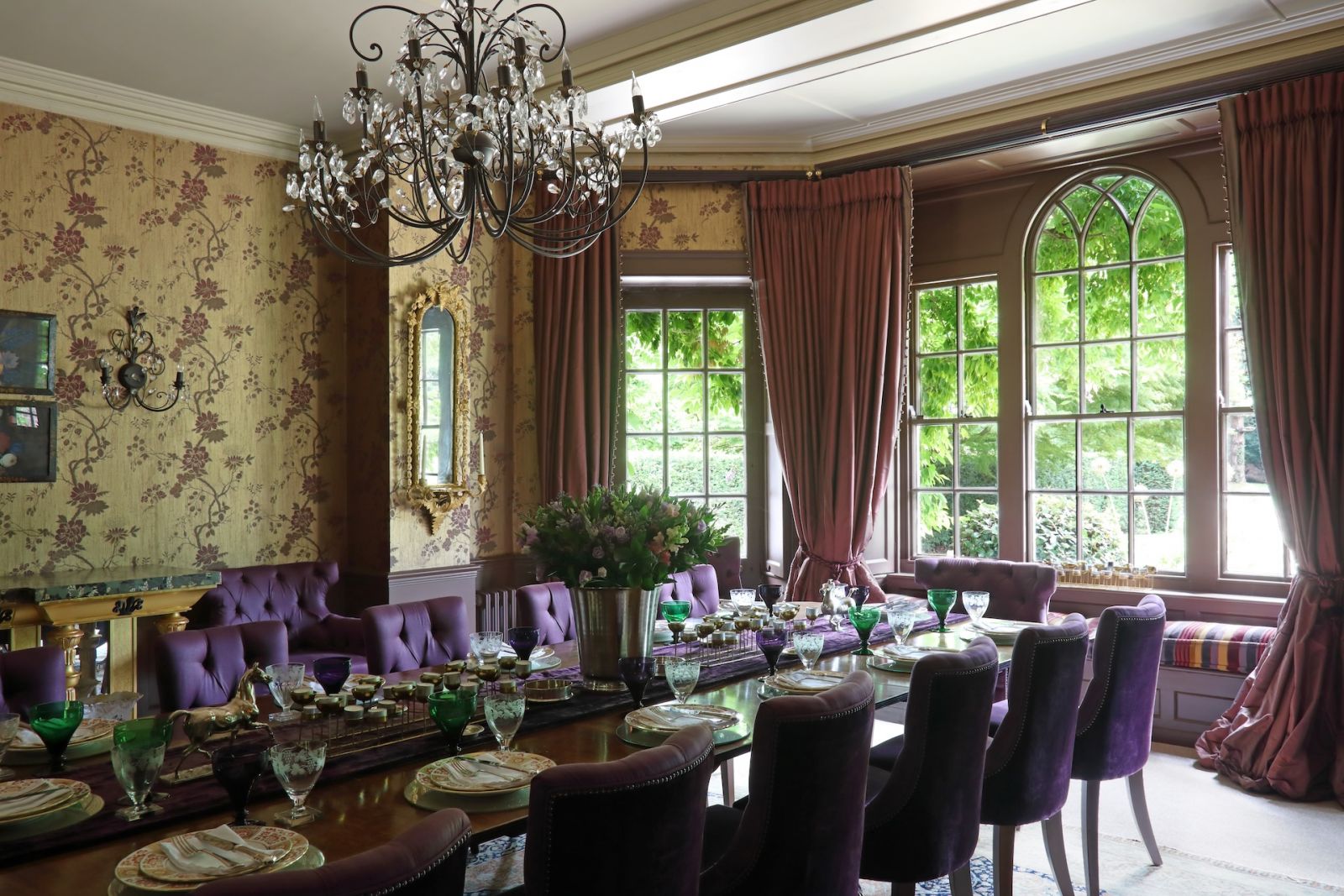 The formal dining room with Zoffany wallpaper and dado panelling in a deep aubergine eggshell finish; the bay window has seating framed by rose taffeta curtains with gold fringe, matching the crystal antique pendant that hangs above the mahogany dining table with Robert Langford button-backed dining chairs