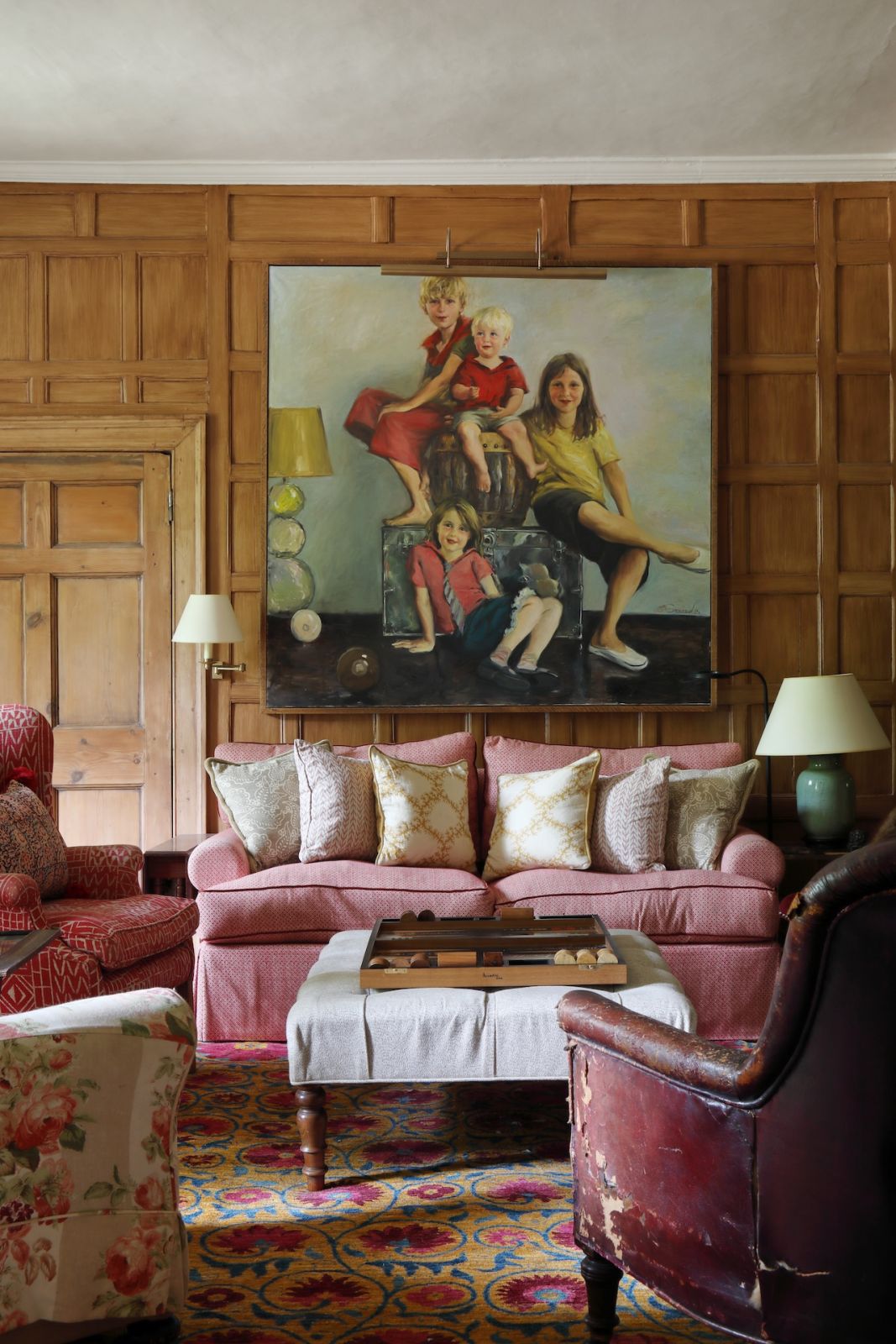 The library snug in this Sussex, England home, with antique oak panelled walls with natural hessian; it also features a Pomegranate rug from The Rug Company that coordinates with a Paolo Moschino herringbone sofa piped in red velvet and a vintage leather armchair