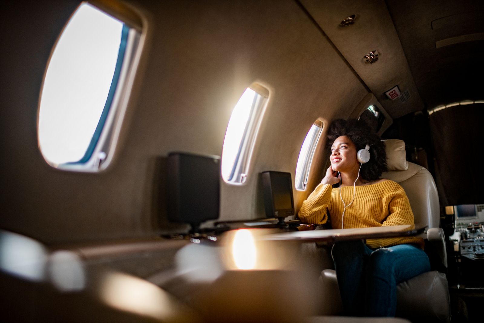 Young fashionable woman sitting on a private airplane and looking through a window while listening to music through headphones