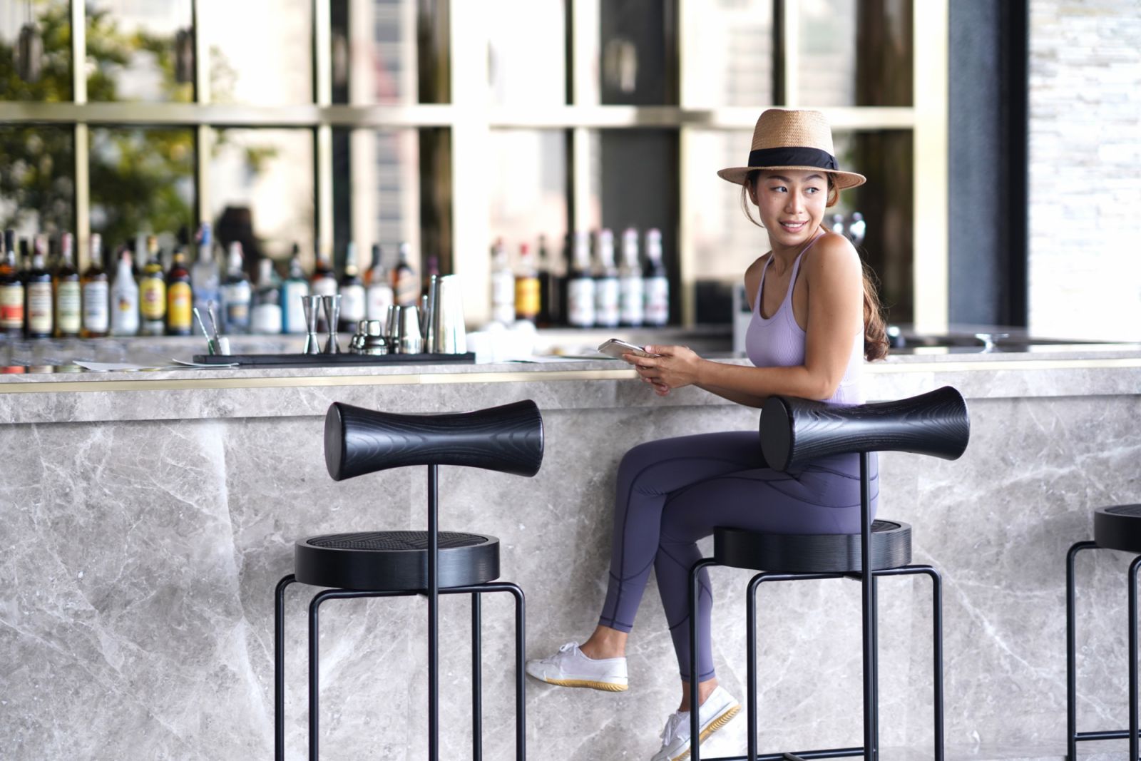 Fashionable young Asian woman sitting on bar stool at hotel coffee shop looking away and smiling while hand holding mobile phone