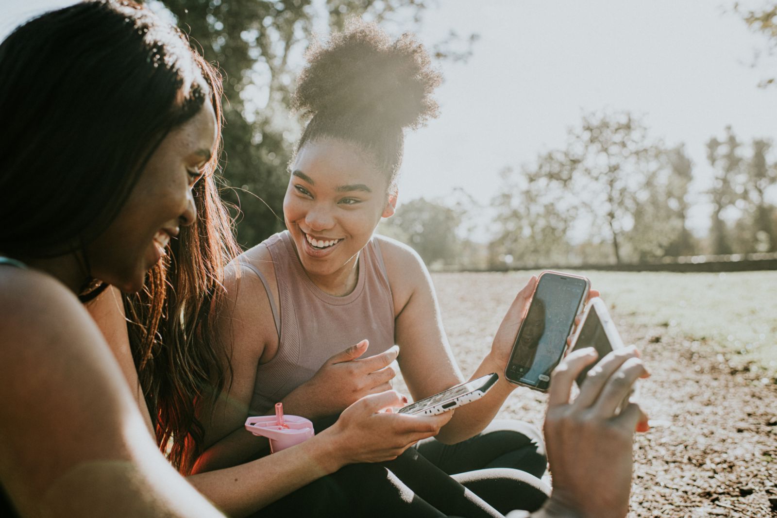 Three healthy young girls in sportswear sit outdoors in the sun wearing sportswear. One holds her mobile phone up to another girl who laughs. The scene is relaxed and happy. Conceptual with space for copy.