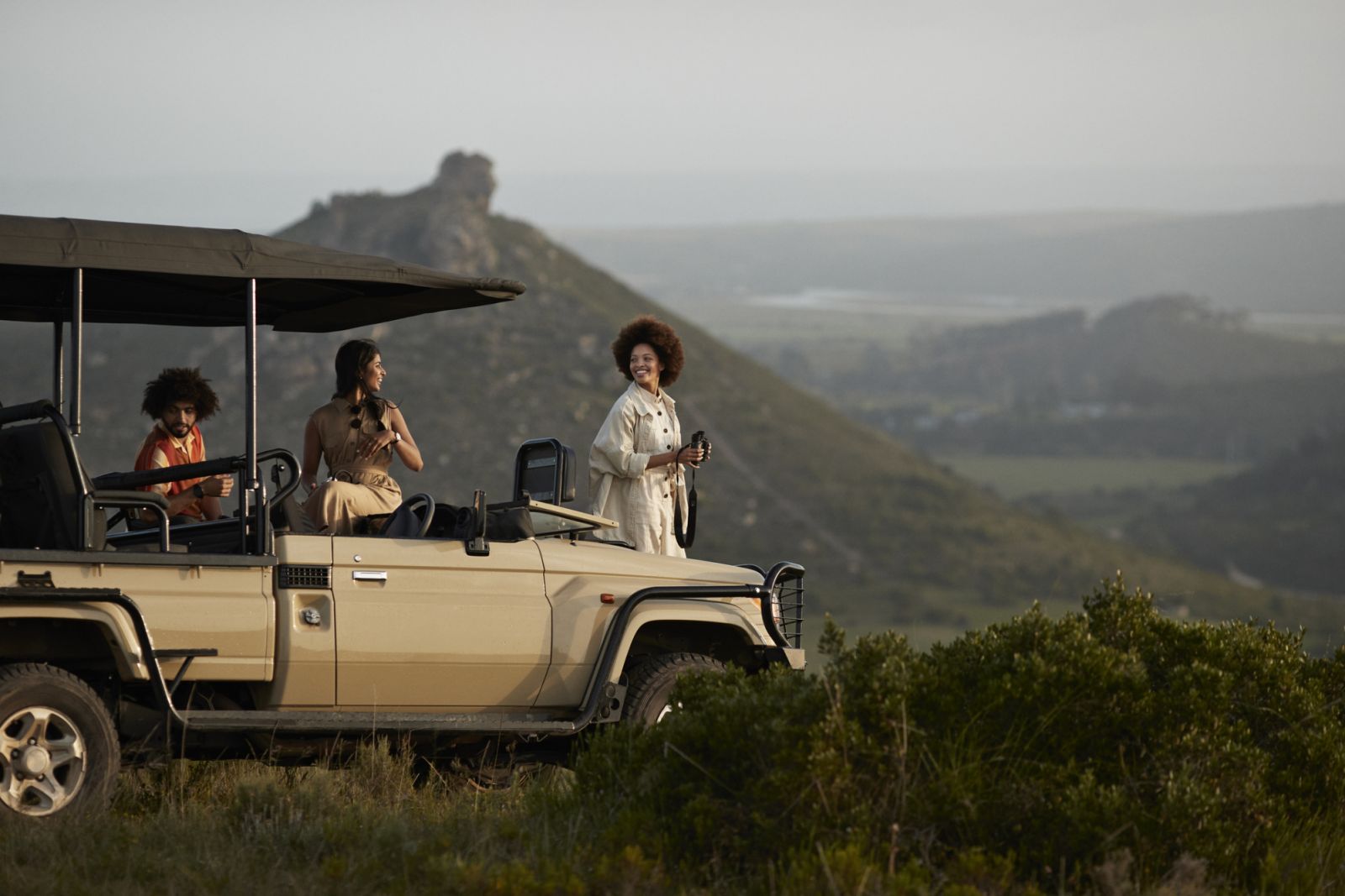 Smiling young woman with binoculars standing near friends in safari vehicle in wildlife park against sky during sunset