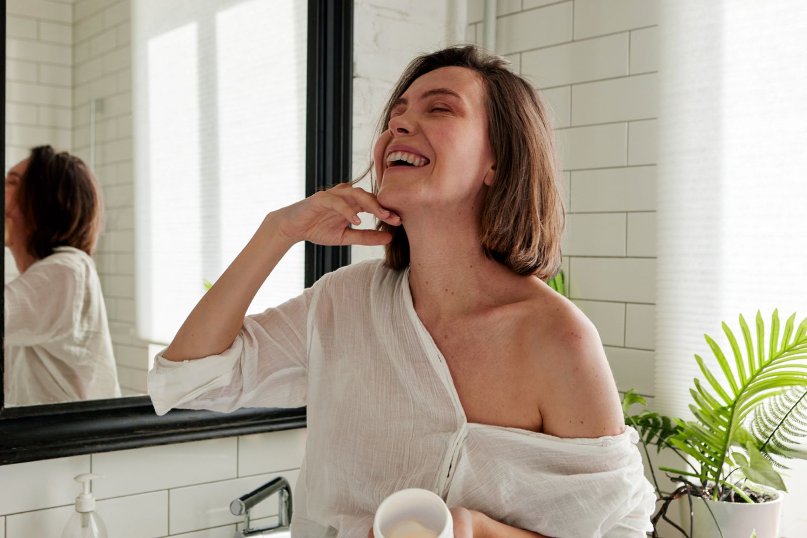 Portrait of positive lady applying cream on face standing with closed eyes in cozy bathroom. She gesticulating hands stroking her chin. Treatment procedure concept