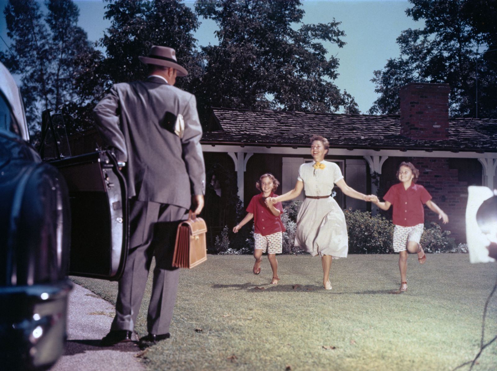 A woman and two girls hold hands as they run across a lawn to greet a man getting out of a car with a briefcase in his hand and a newspaper under his arm, 1950s. (Photo by FPG/Getty Images)