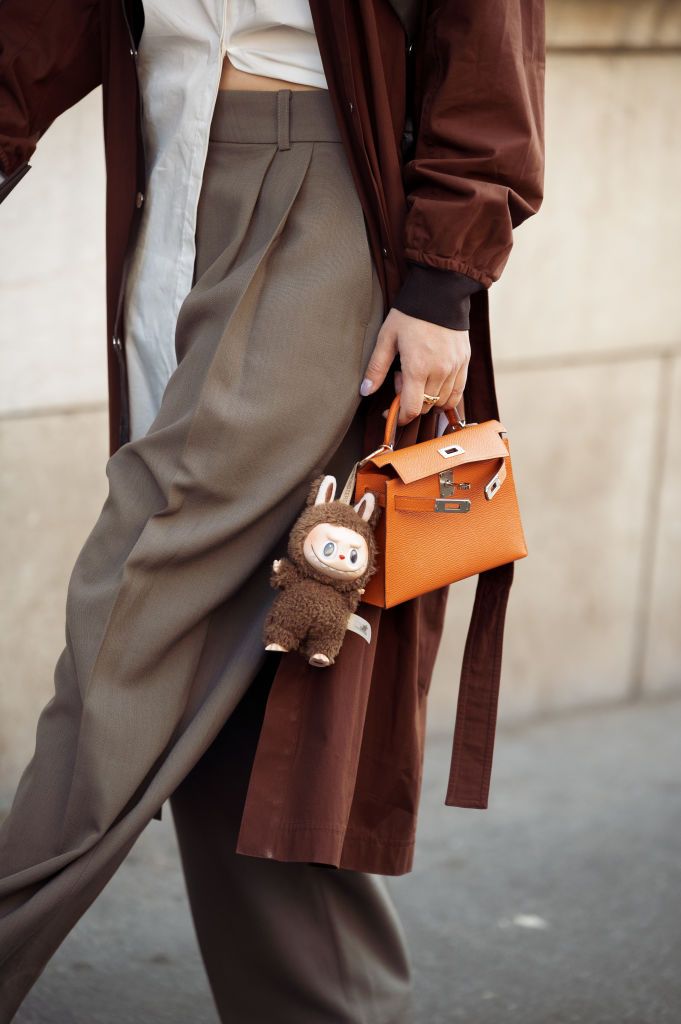 PARIS, FRANCE - MARCH 08: A guest wears brown coat, white blouse, orange Hermes bag with a bag charm brown Labubu monster, grey pants outside the Hermes fashion show during the Womenswear Fall/Winter 2025/2026 as part of Paris Fashion Week on March 08, 2025 in Paris, France. (Photo by Raimonda Kulikauskiene/Getty Images)