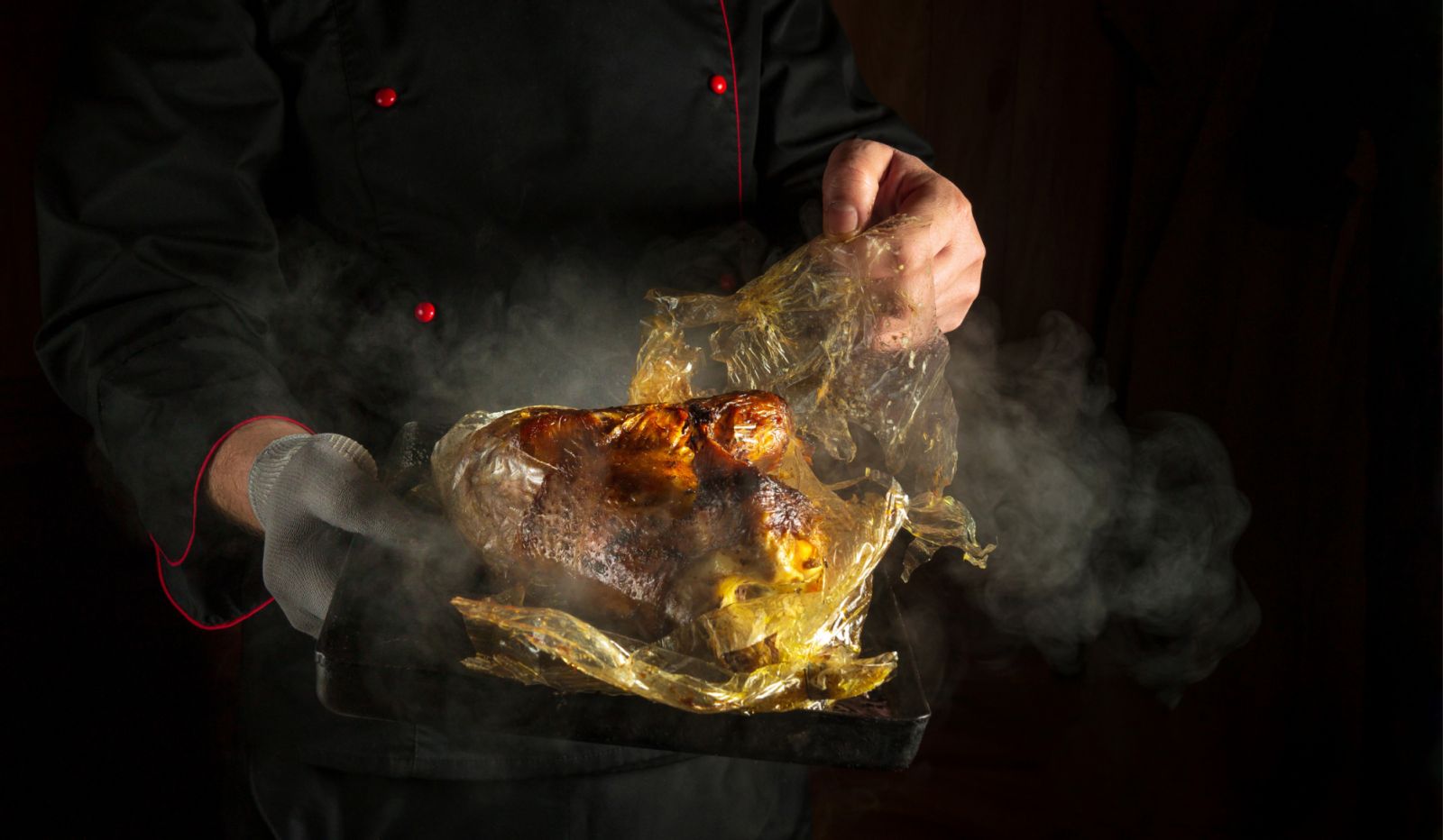Cooking baked chicken on a baking sheet in the oven. Low key concept of roasting a bird for dinner with dark space for advertising. Chef in black uniform with tray.