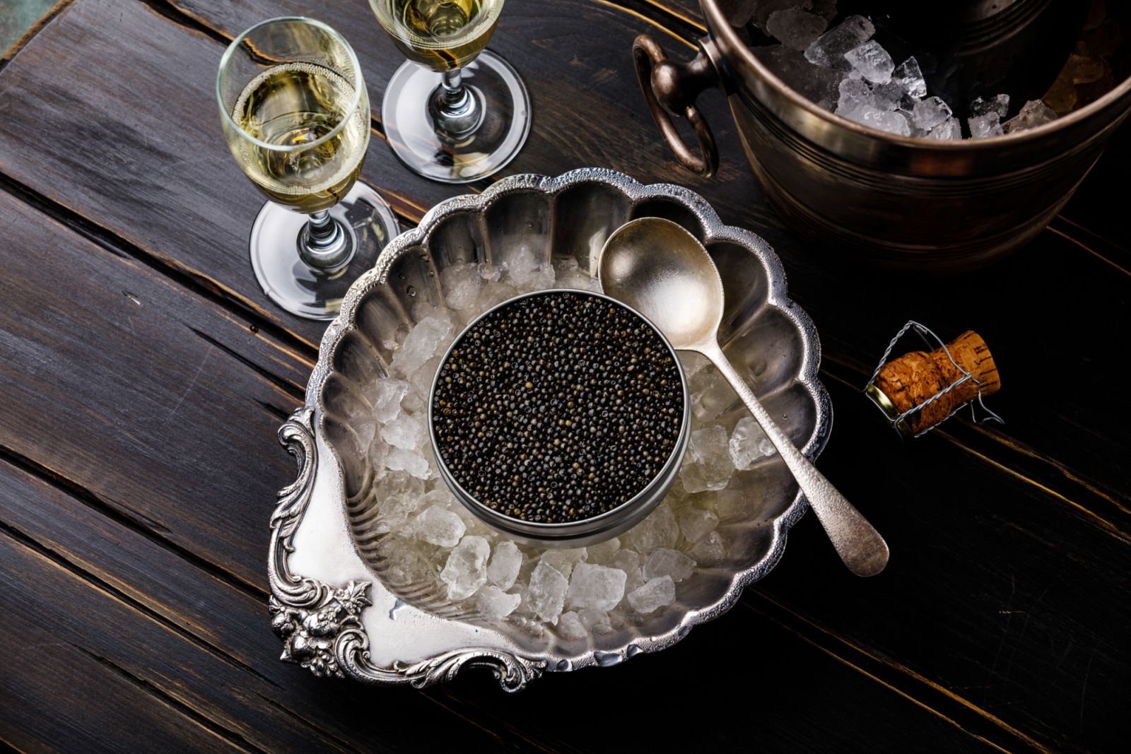 Black caviar in can on ice in silver bowl and champagne on black wooden background