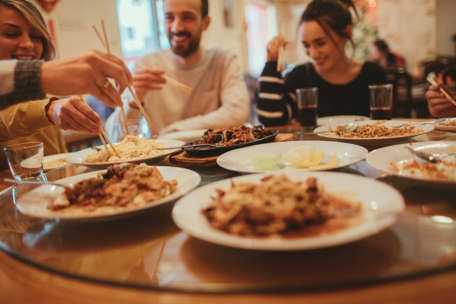 Photo of a group of friends having fun while eating with sticks in a Chinese restaurant. Food is in a foreground, while people are in background.