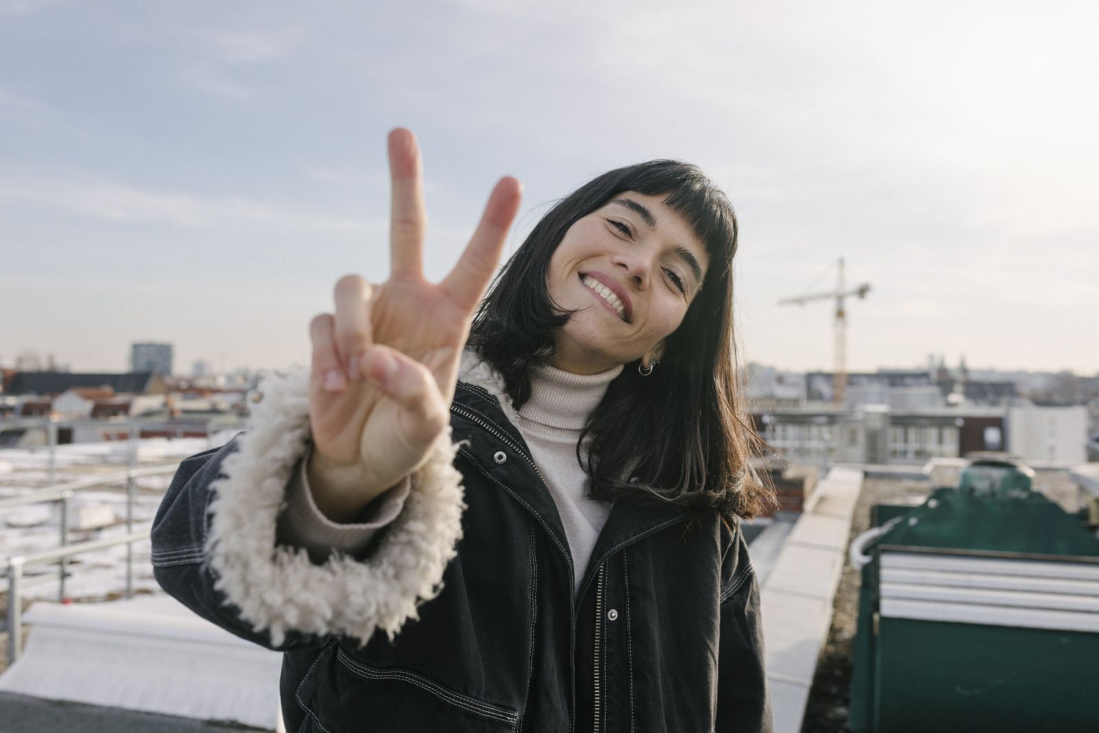 A portrait of a woman wearing a warm jacket and standing on a rooftop while giving a peace sign.