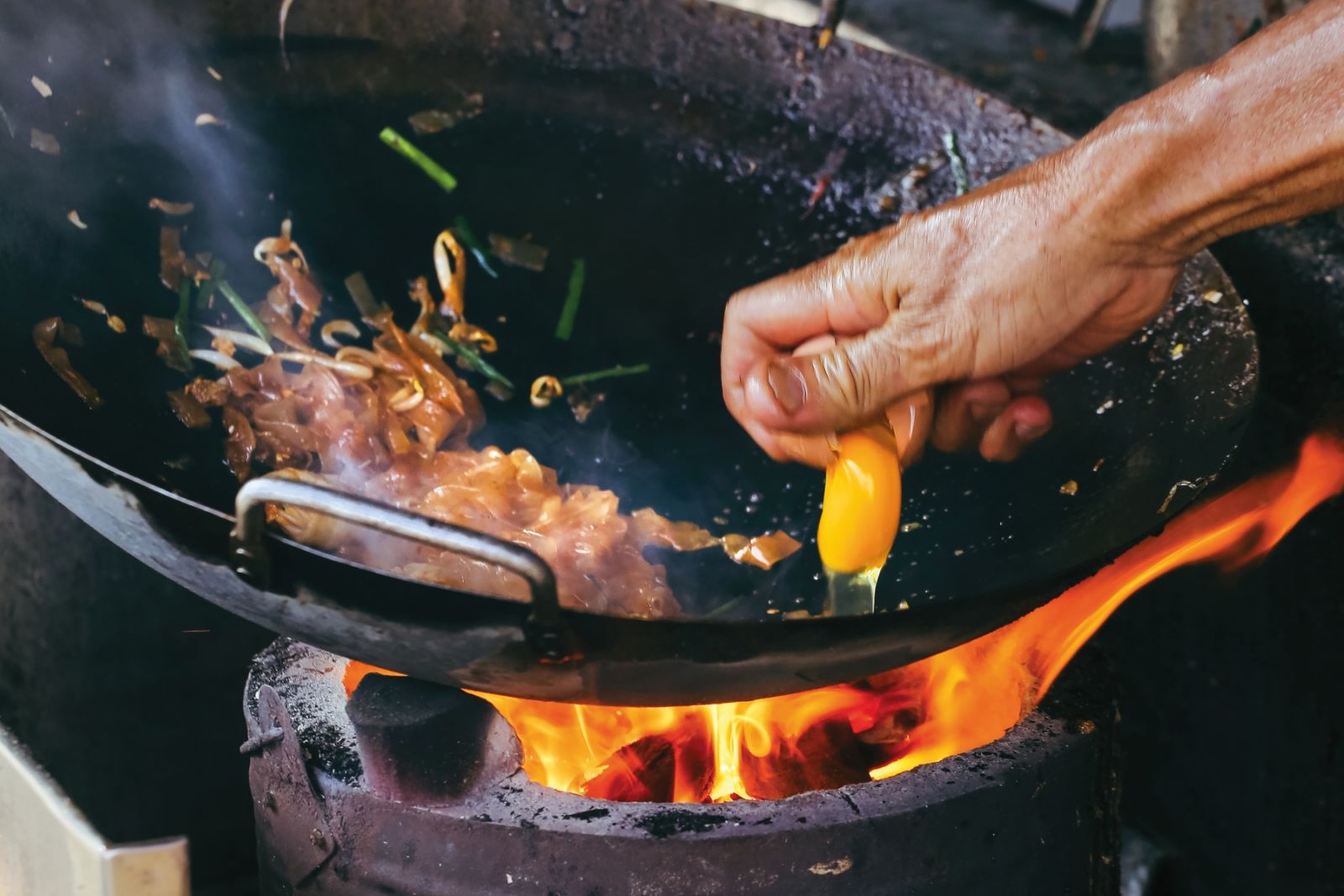 High angle close up view of male hand breaking a chicken egg onto the frying pan of  char kway teow  - Malaysian stir fried flat rice noodles - with chicken egg, prawns, and lap cheong (Chinese sausage), George Town, Penang island, Malaysia