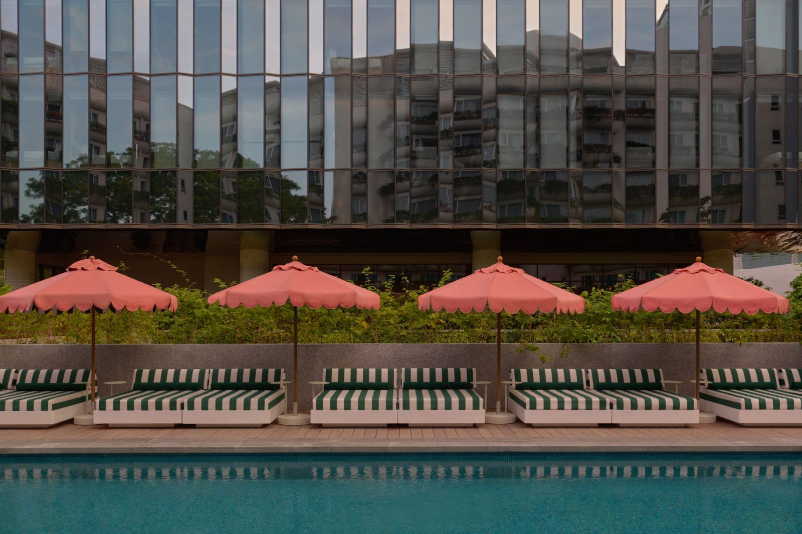 Poolside loungers at The Standard Singapore with distinctive coral umbrellas and green striped cushions against a reflective glass façade—a playful reinterpretation of classic resort aesthetics (Photo: The Standard Singapore)