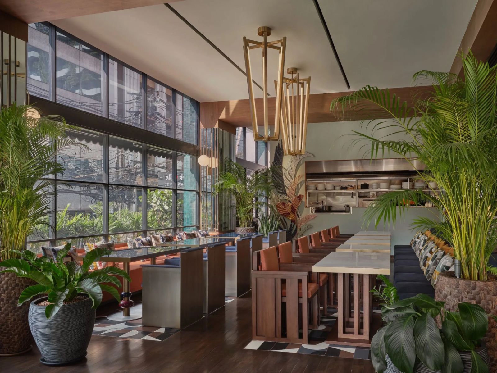 Restaurant at Public House Hotel Bangkok featuring brass geometric pendant lighting, abundant tropical foliage, and wooden slatted chairs set against floor-to-ceiling windows—blending indoor-outdoor dining with mid-century modern design elements and a glimpse of Bangkok's urban landscape beyond (Photo: Public House Hotel Bangkok)
