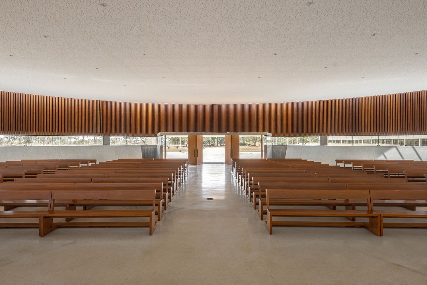 Interior of the circular nave with wooden pews arranged to focus on the altar, showing the perimeter clerestory window and wooden slat walls that warm the concrete space (Photo: ARQBR Arquitetura e Urbanismo)