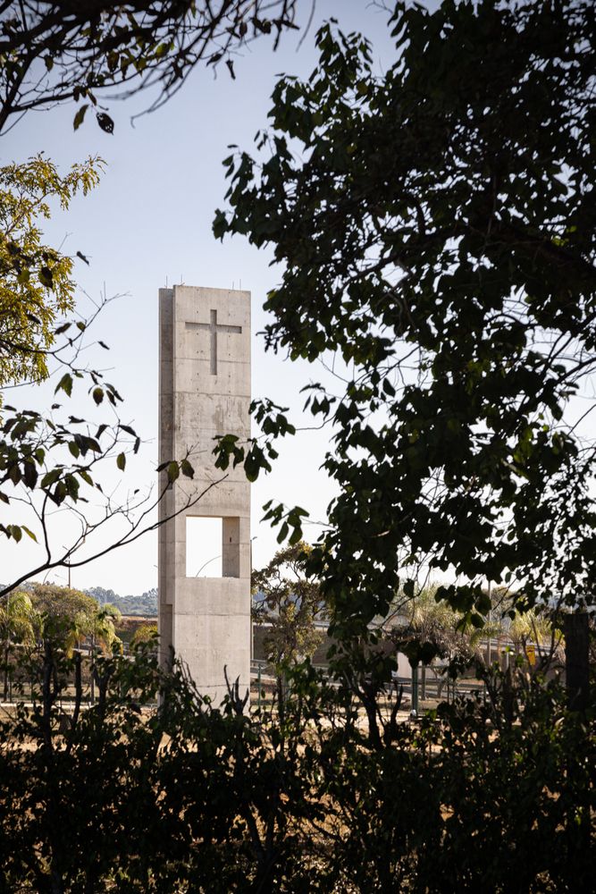 The bell tower framed through native vegetation, showing how the concrete structure with its cruciform cutout relates to Brasilia's natural context (Photo: ARQBR Arquitetura e Urbanismo)
