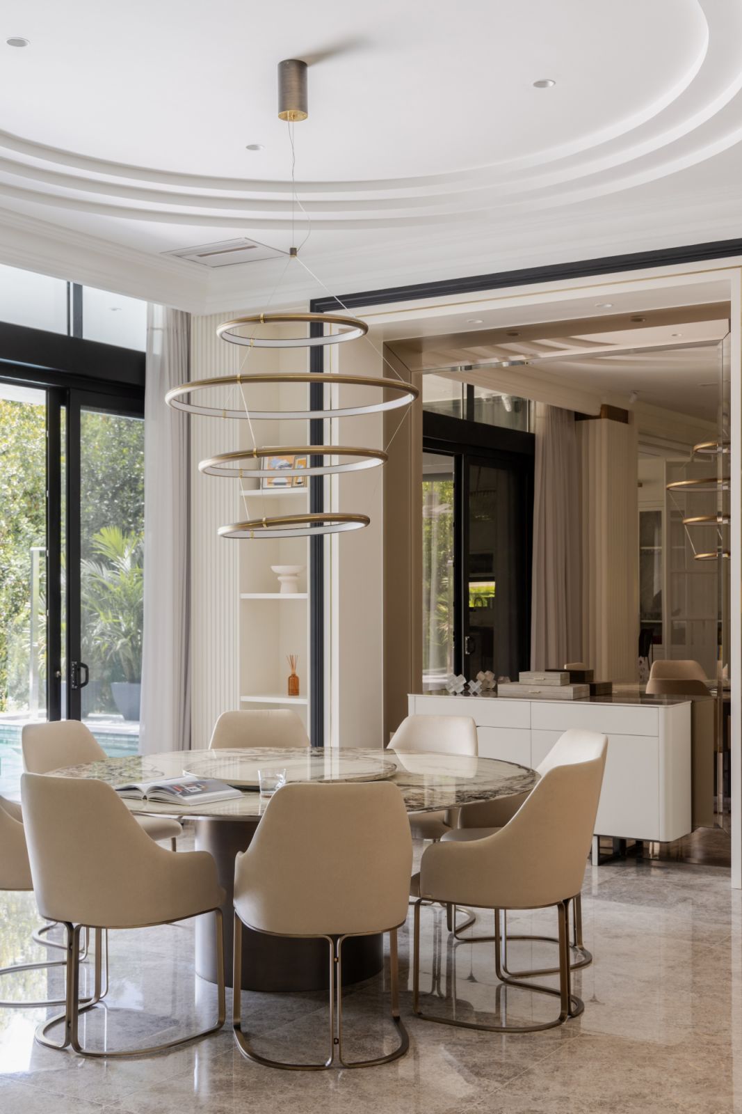 Contemporary dining space with marble tabletop, designer seating, and tiered circular chandelier in neutral tones with natural light