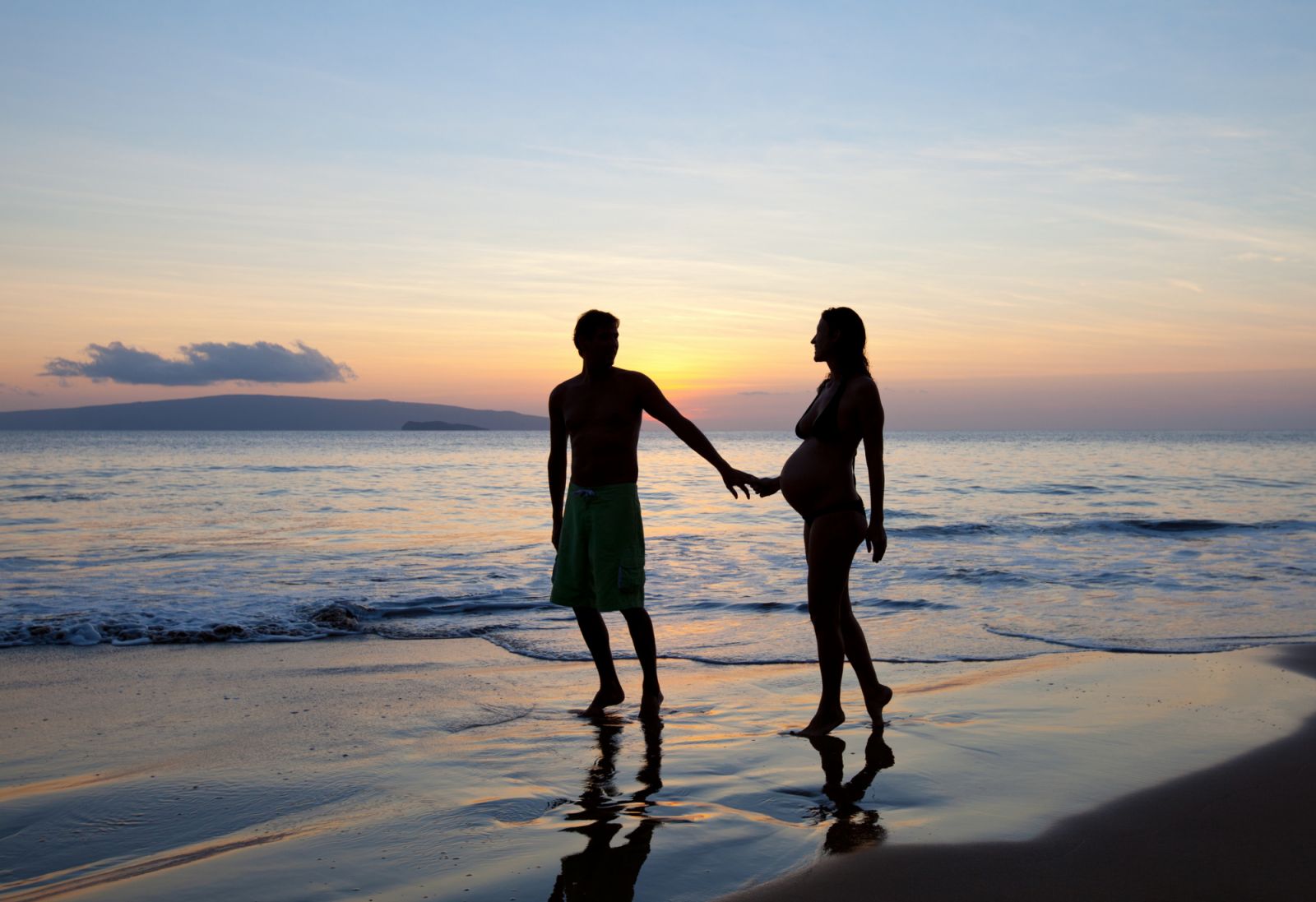Silhouette romantic pregnant couple at tropical beach sunset in Maui, Hawaii.