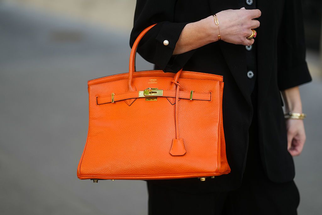 PARIS, FRANCE - APRIL 09: An orange Hermes Birkin bag in grained leather with structured handles, gold-toned hardware, a signature front lock, and tonal stitching is carried. A black blazer with pearl button cuffs is worn. A gold cuff bracelet and a sculptural gold ring with red enamel detailing complete the look. Detail shot, during a street style fashion photo session, on April 09, 2025 in Paris, France. (Photo by Edward Berthelot/Getty Images)