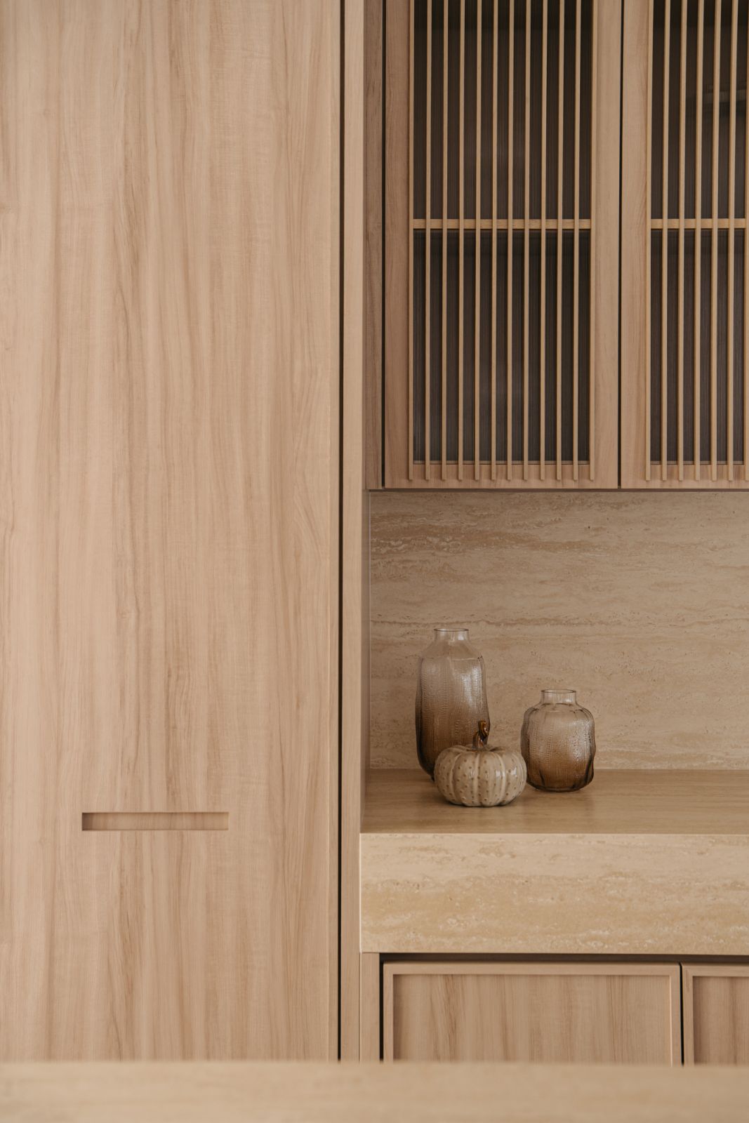 Bespoke kitchen cabinetry featuring slatted wood details, integrated recessed handles and artisanal vessels against travertine backsplash