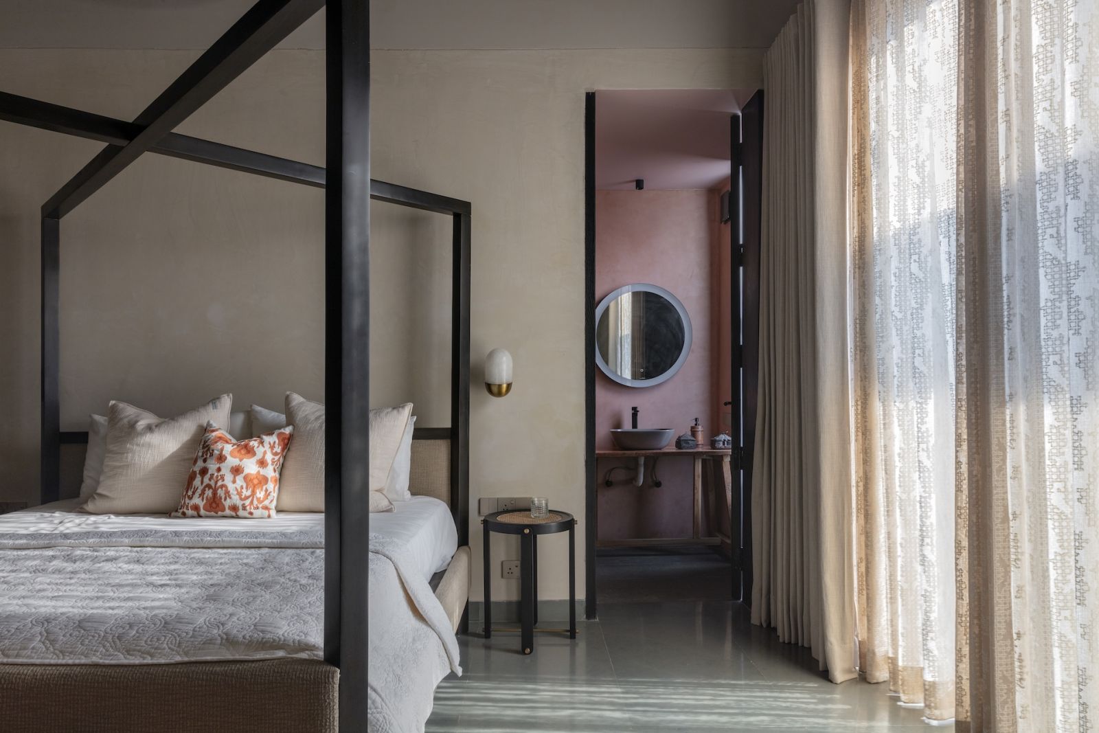 The guest bedroom in a Hyderabad, India home with an X-frame bed, offering glimpses of salmon hues in the ensuite bathroom