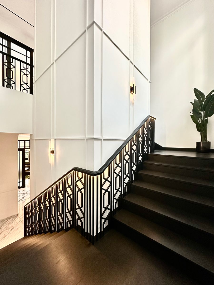 The dark, art deco-inspired staircase and balustrade, paired with the white wainscoted walls, evoke a sense of colonial splendour