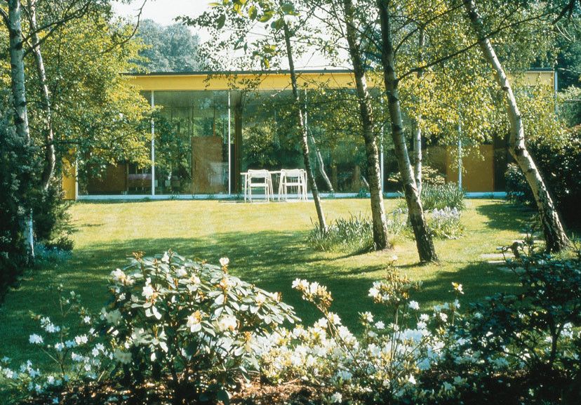 Exterior garden view of Richard Rogers’ 22 Parkside House in Wimbledon with a modernist glass façade and yellow steel frame blending harmoniously with the lush landscape (Photo: Courtesy of Rogers Stirk Harbour + Partners LLP)
