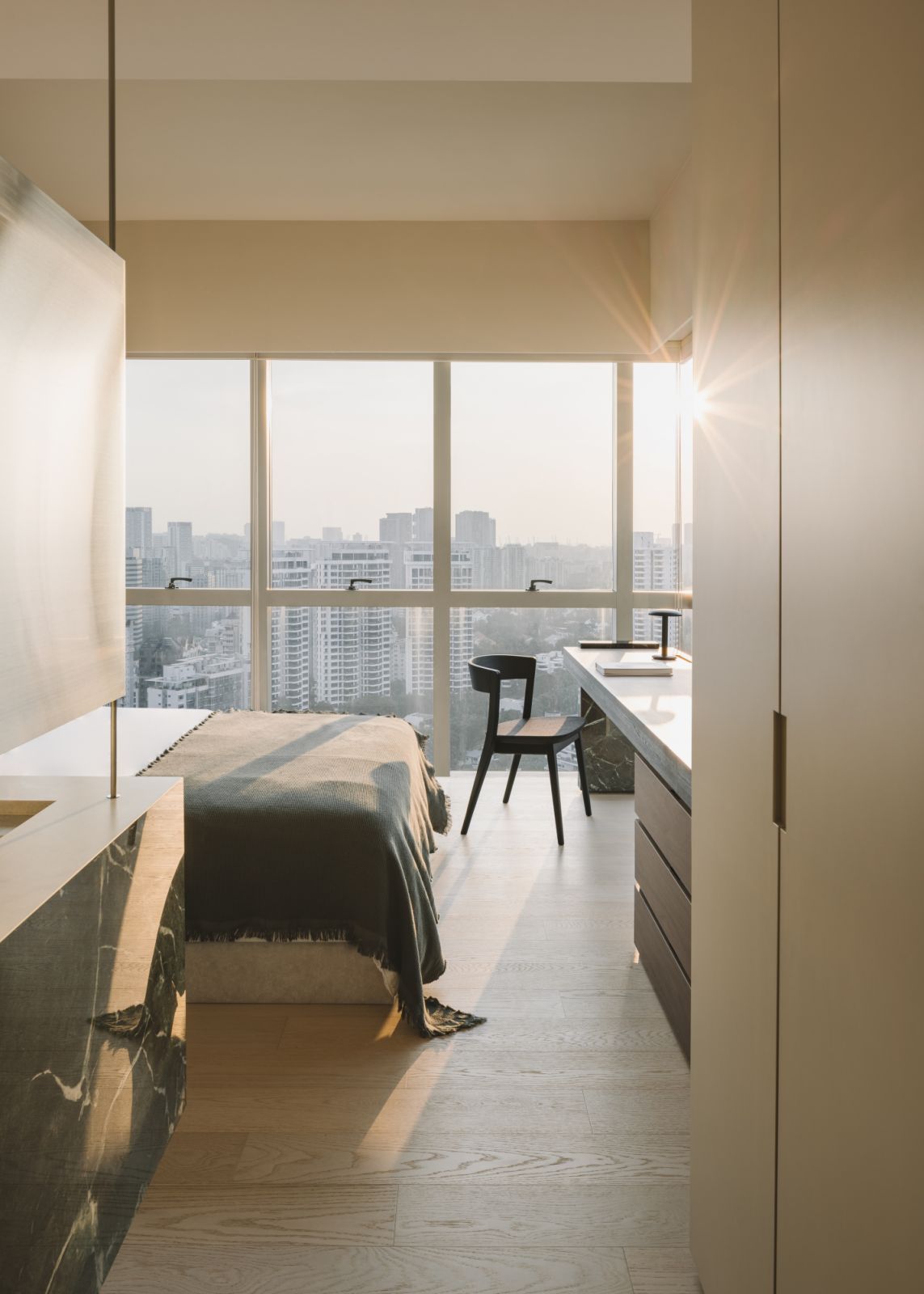 Framed by a full-height wall of glazing, the bedroom reveals panoramic views of Singapore’s skyline—contrasted by the quiet tactility of wood, stone, and wool