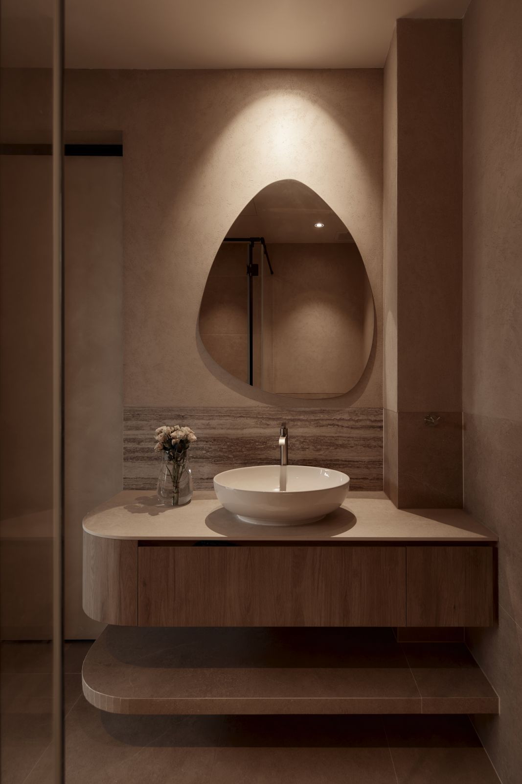 A softly lit asymmetrical mirror adds visual intrigue to the powder room, enhanced by curved joinery and tactile finishes