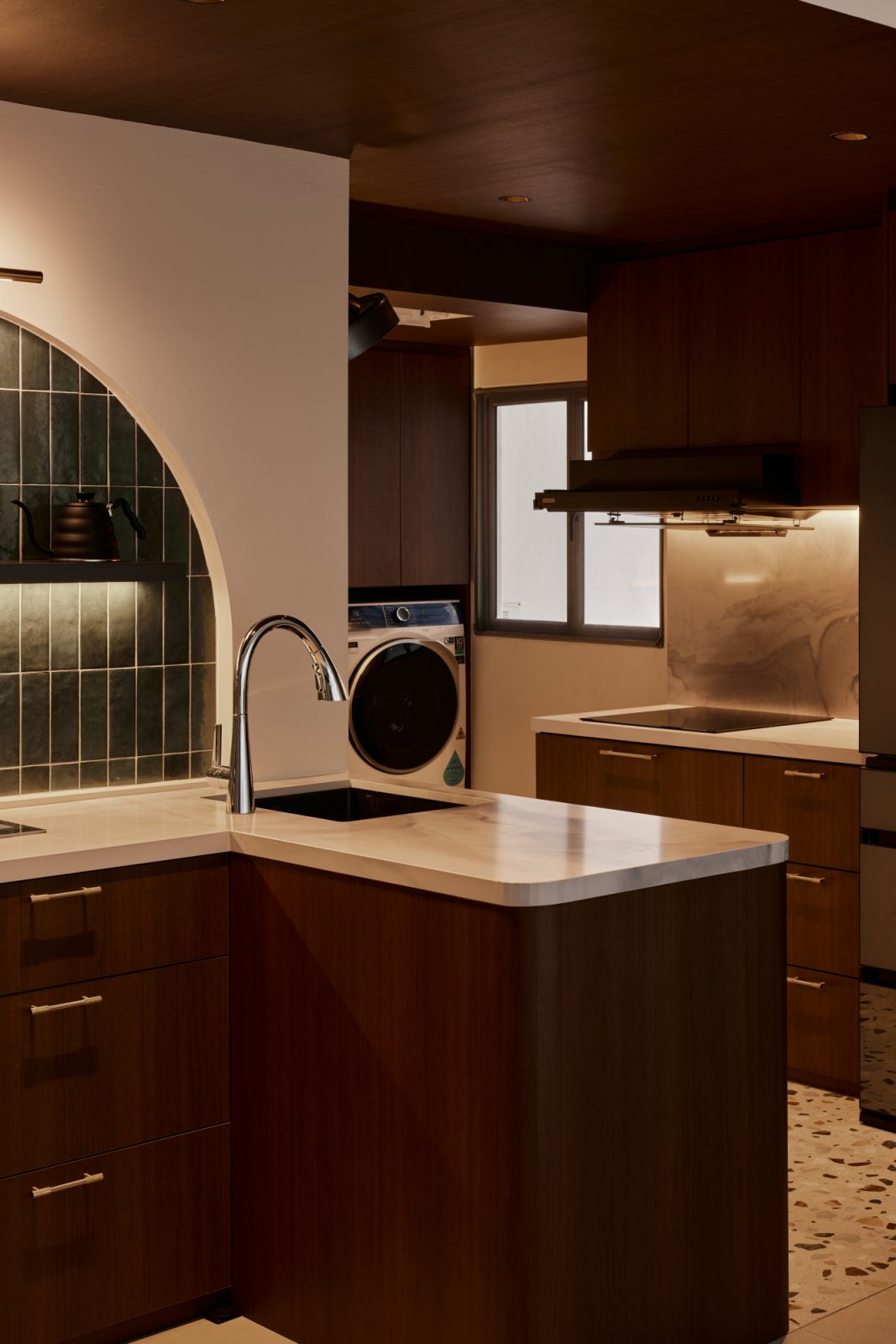 The transition from day to night subtly shifts the kitchen’s mood, enhanced by warm lighting and rich materiality