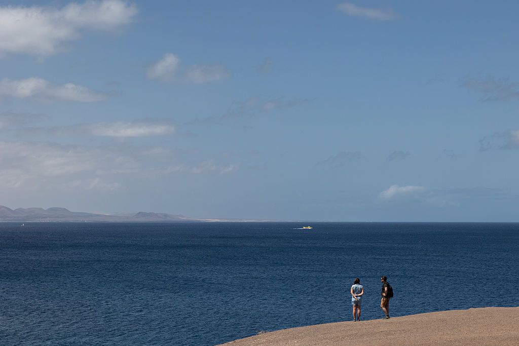LOS AJACHES, SPAIN - APRIL 22: A woman and a man stand on a cliff overlooking the Atlantic Ocean and the island of Fuerteventura on April 22, 2025 in Lanzarote, Spain. Lanzarote, a Spanish Canary Island and a designated UNESCO Biosphere Reserve, lies off the coast of West Africa, famous for its unique volcanic landscapes, stunning beaches, and a year-round warm climate that draws tourists to its natural beauty and rich cultural identity. (Photo by Emanuele Cremaschi/Getty Images)