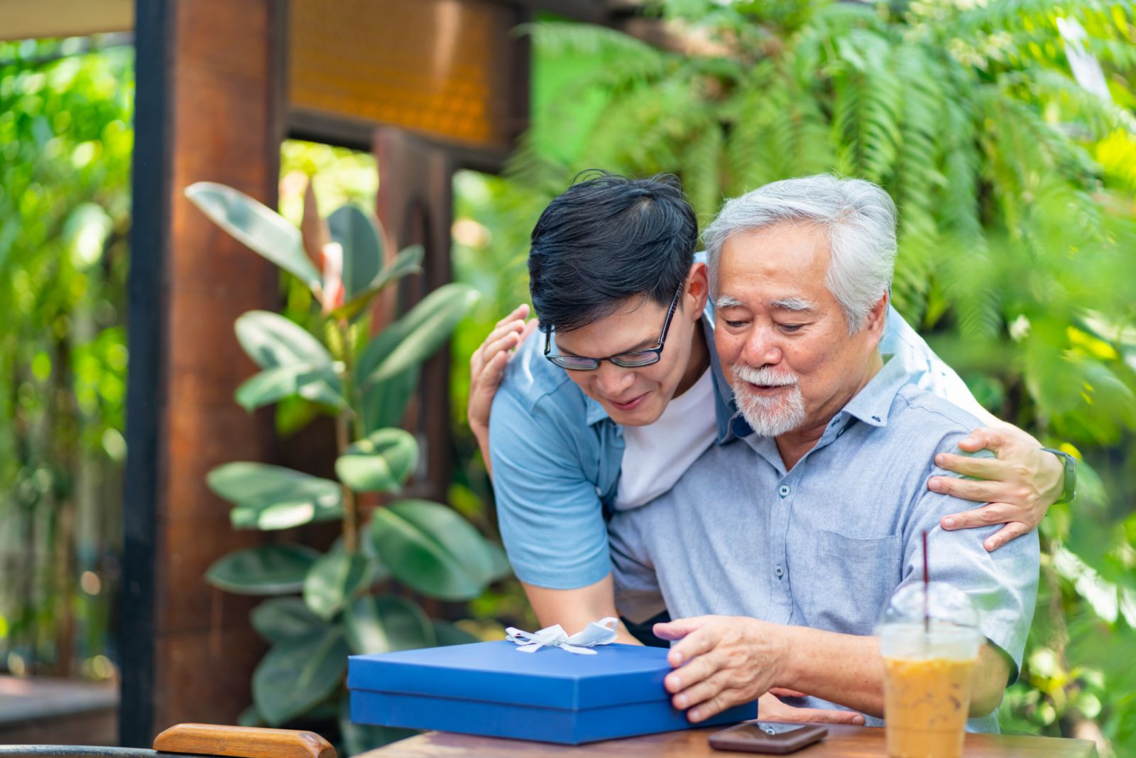 Asian man surprise hugging elderly father from back at outdoor garden cafe restaurant on summer vacation. Family relationship, holiday celebrating, father's day and old people health care concept