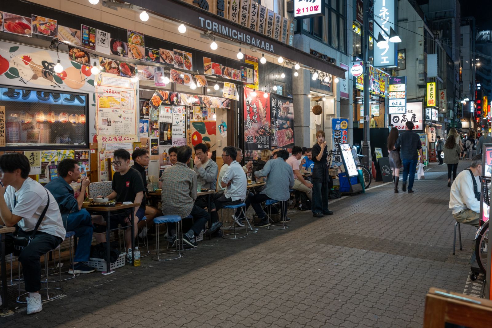 13 October 2024, Tokyo, Japan: Crowded outdoor eatery with diners at tables, colorful signage, and busy street atmosphere