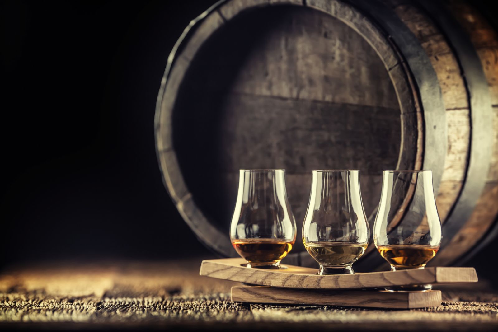 Glencairn whiskey tasting cups on a wooden serving, with a whisky barrel in the dark background.