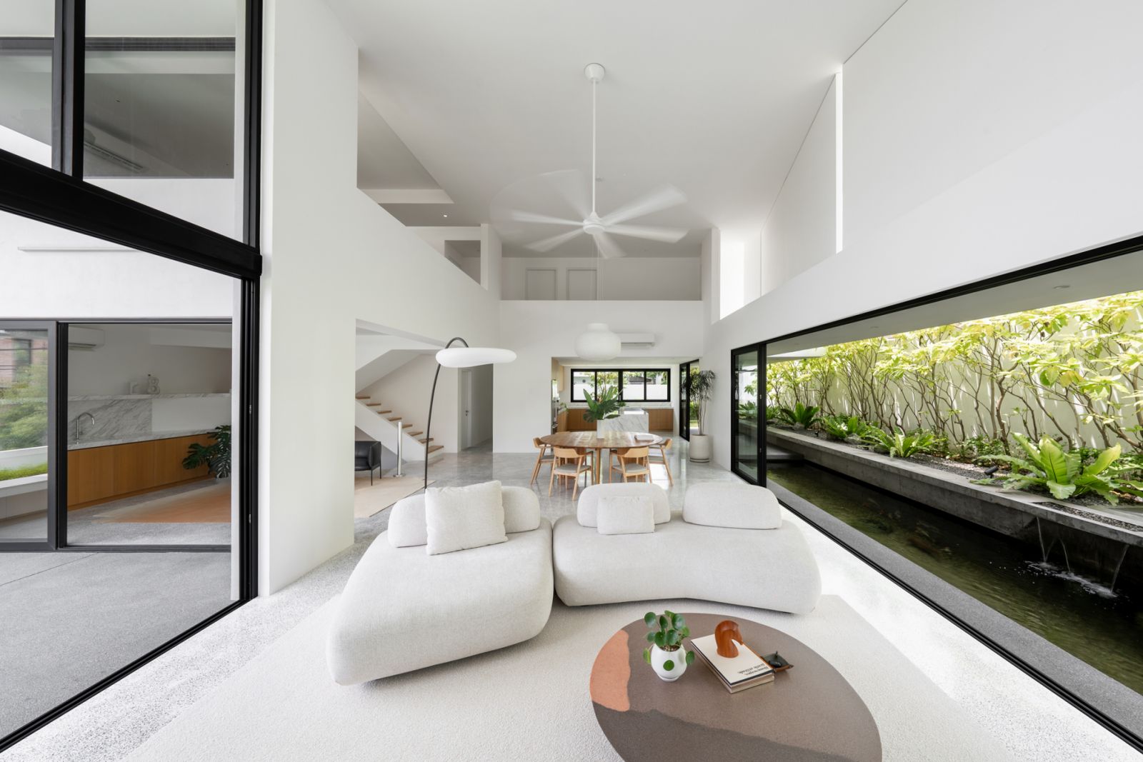 Double-height living space in Malaysian bungalow features contemporary furniture, terrazzo flooring, and panoramic koi pond views through floor-to-ceiling glazing