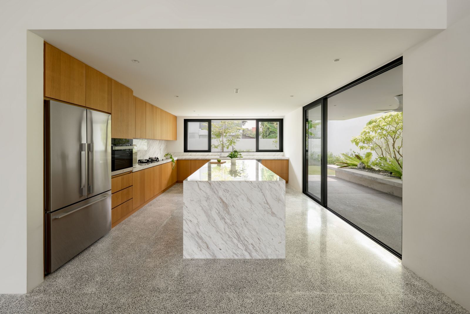 Modern bungalow kitchen renovation showcases marble waterfall island, warm oak timber cabinets, and seamless terrazzo flooring connecting to outdoor garden space