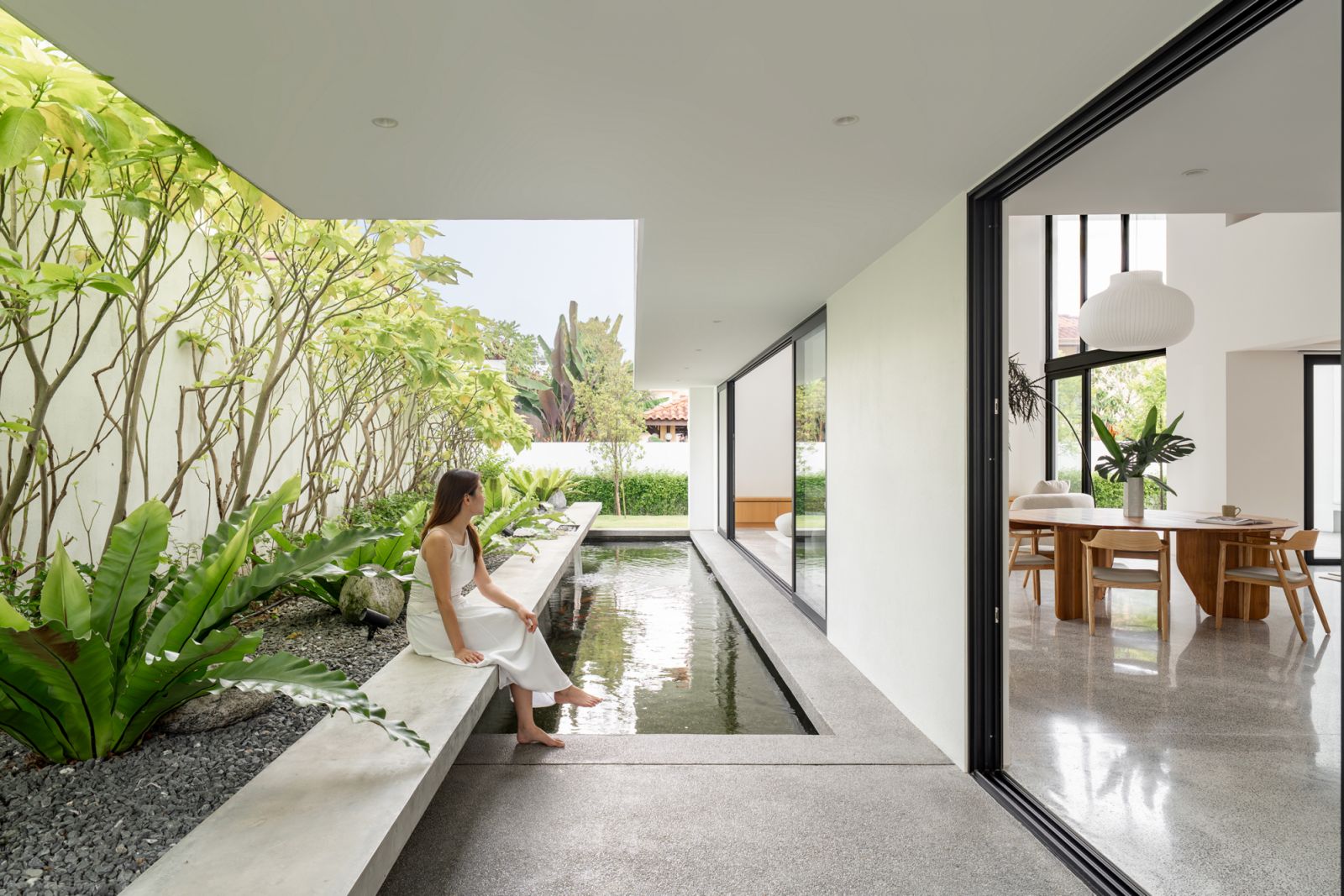 Covered outdoor terrace of Malaysian bungalow renovation features integrated koi pond with elevated planters, shade trees, and seamless indoor-outdoor living connection