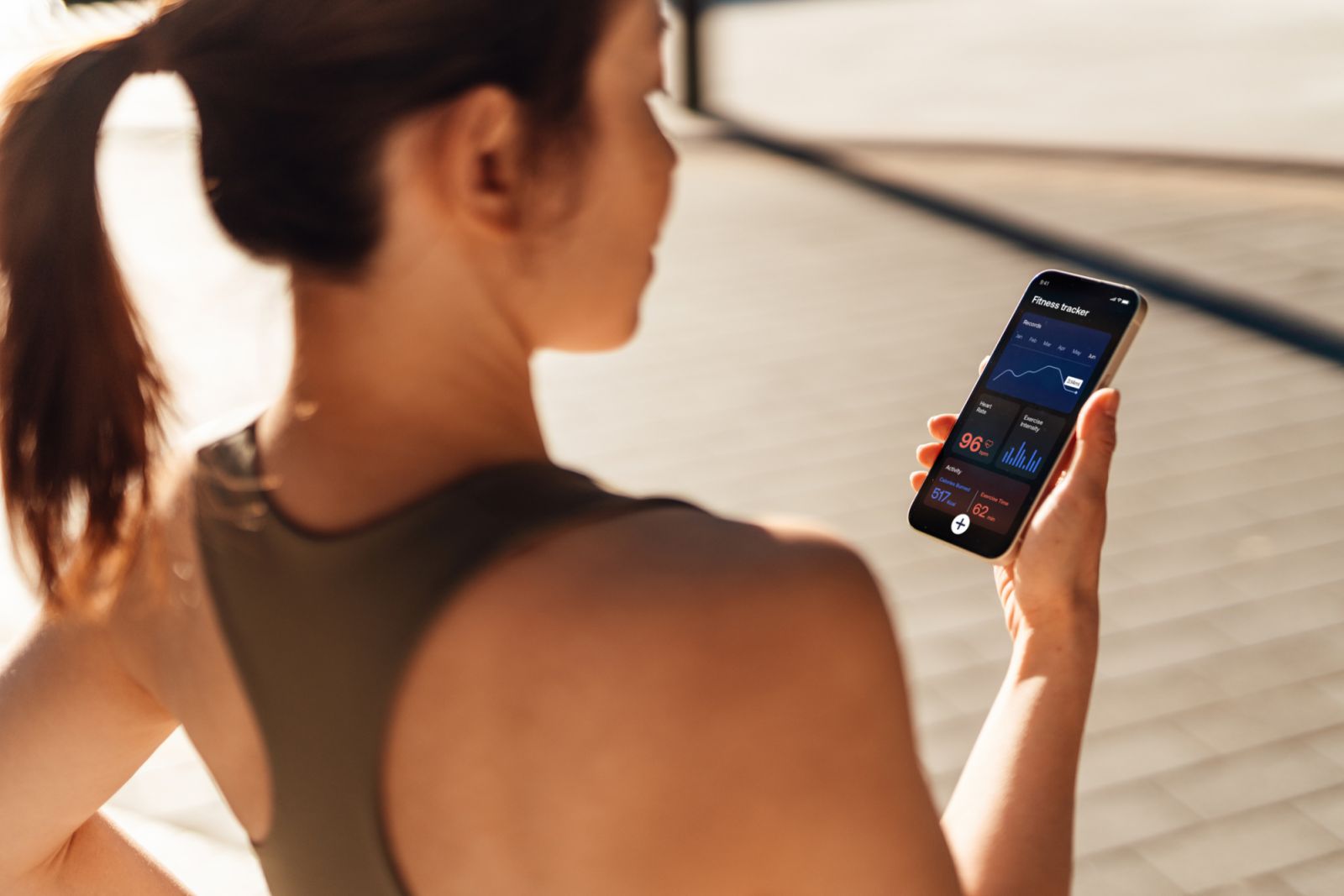 Over the shoulder view of a sporty woman tracking and analysing her fitness performance with mobile app on smartphone while on a run in the city. FemTech (Female technology).