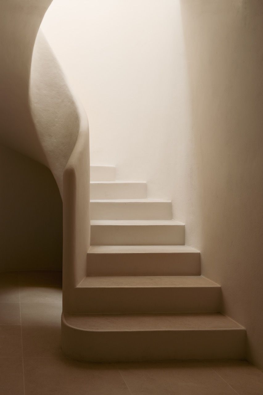 The spiral staircase demonstrates the home’s contemporary interpretation of traditional finca architecture, with curved microcement walls and sandstone flooring creating a sculptural element within the clean interior