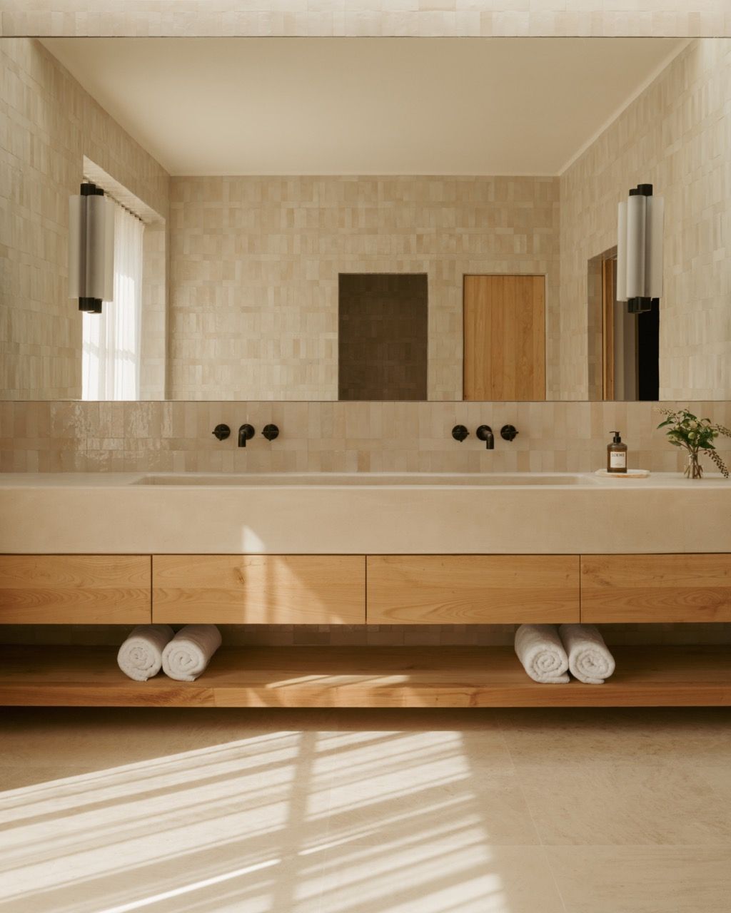 The master bathroom’s double vanity combines limestone surfaces with wooden cabinetry, creating a spa-like atmosphere that reflects the home’s natural material palette