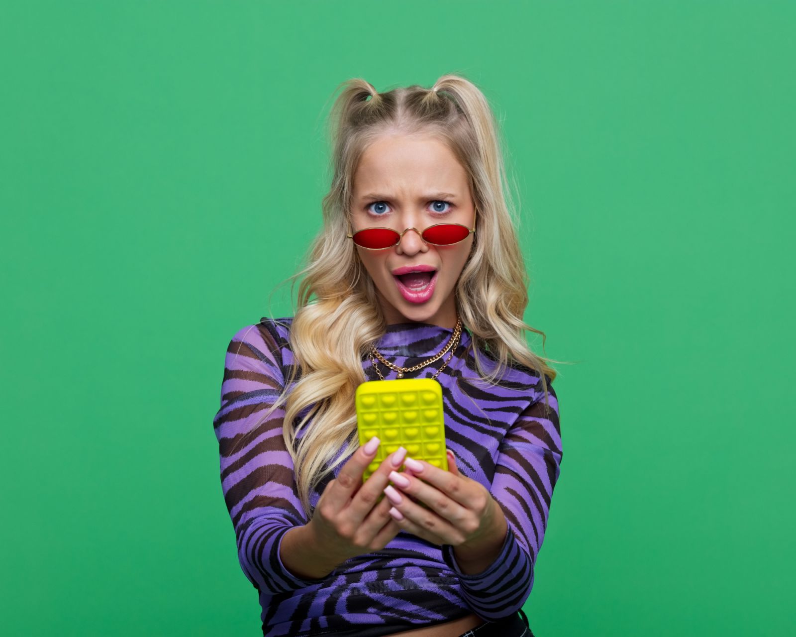 Portrait of shocked young blonde woman wearing purple animal pattern blouse holding mobile phone and looking at camera. Studio shot, green background.