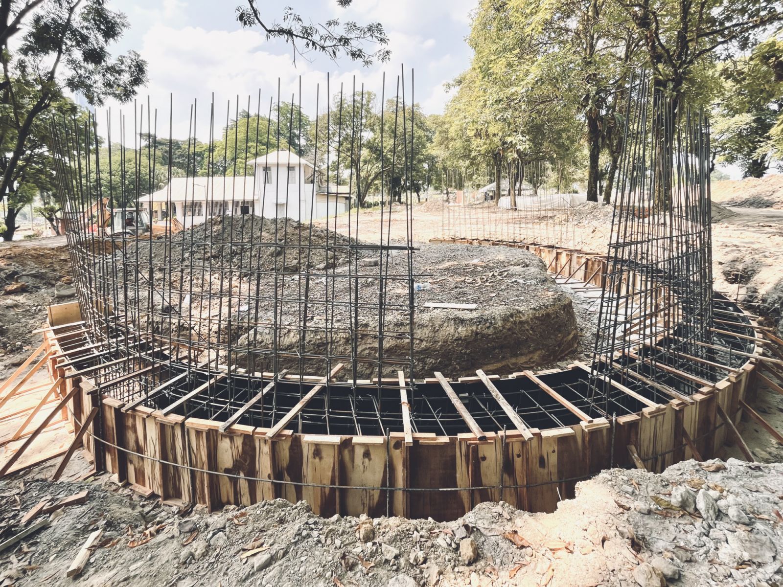Construction phase showing the circular foundation and rebar framework that will support the curved rammed earth walls, preserving the site’s mature trees during building