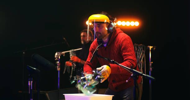 Male sound designer in protective face shield using hammer to break glass jar near microphones and recording soundtrack for movie near colleague in studio