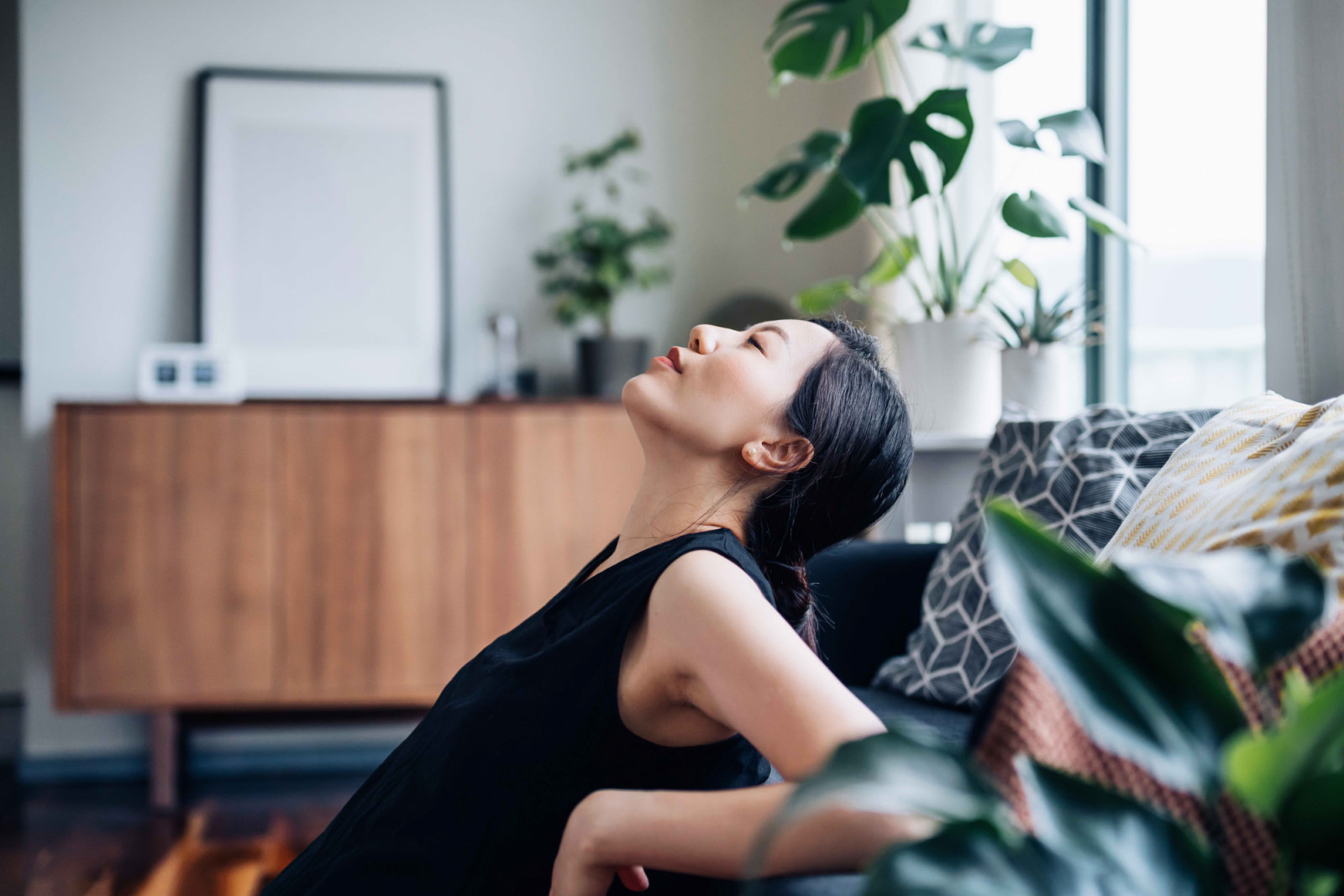 Portrait of a beautiful young Asian woman relaxes by the sofa in the living room, with her eyes closed and head up, enjoying the tranquility during leisure time at cozy home