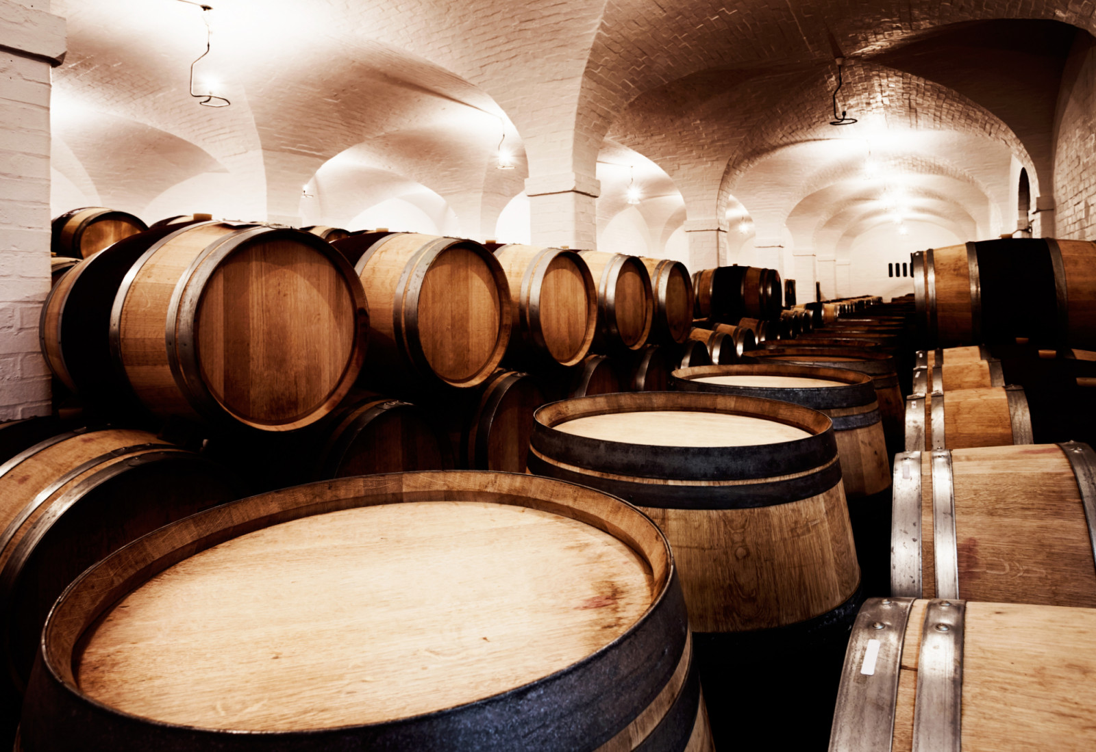 Looking down a long row of stacked wine barrels, used to mature wine, in a winery cellar that resembles the aisle of a chapel, dedicated to wine.