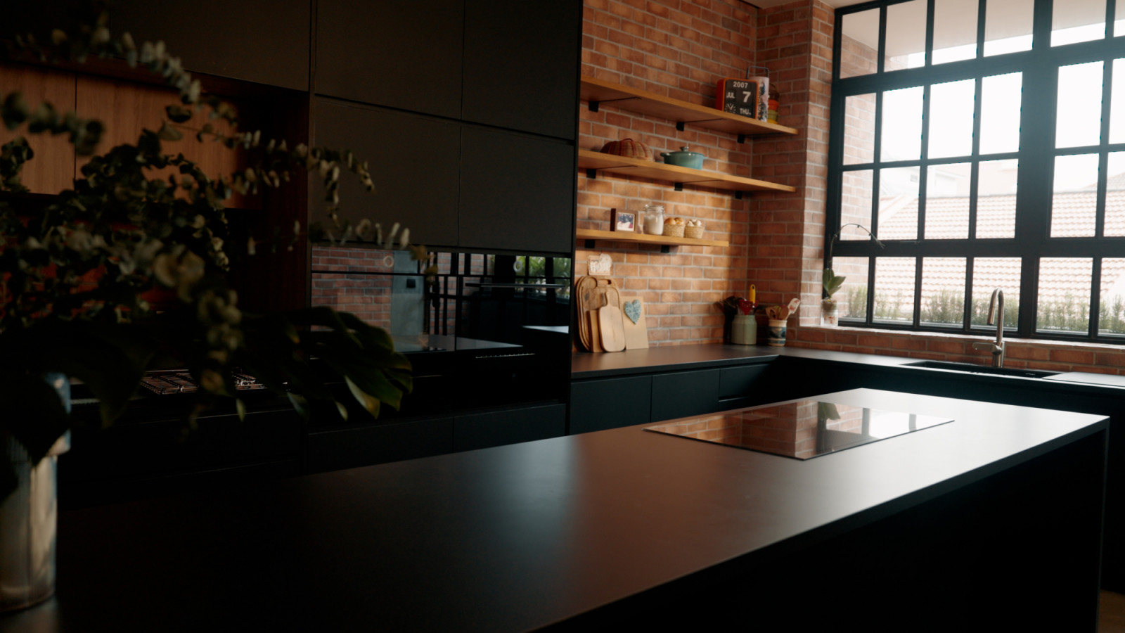 Exposed brickwork and matte black cabinetry create a bold, industrial look in the kitchen, softened by warm wood accents and tropical light