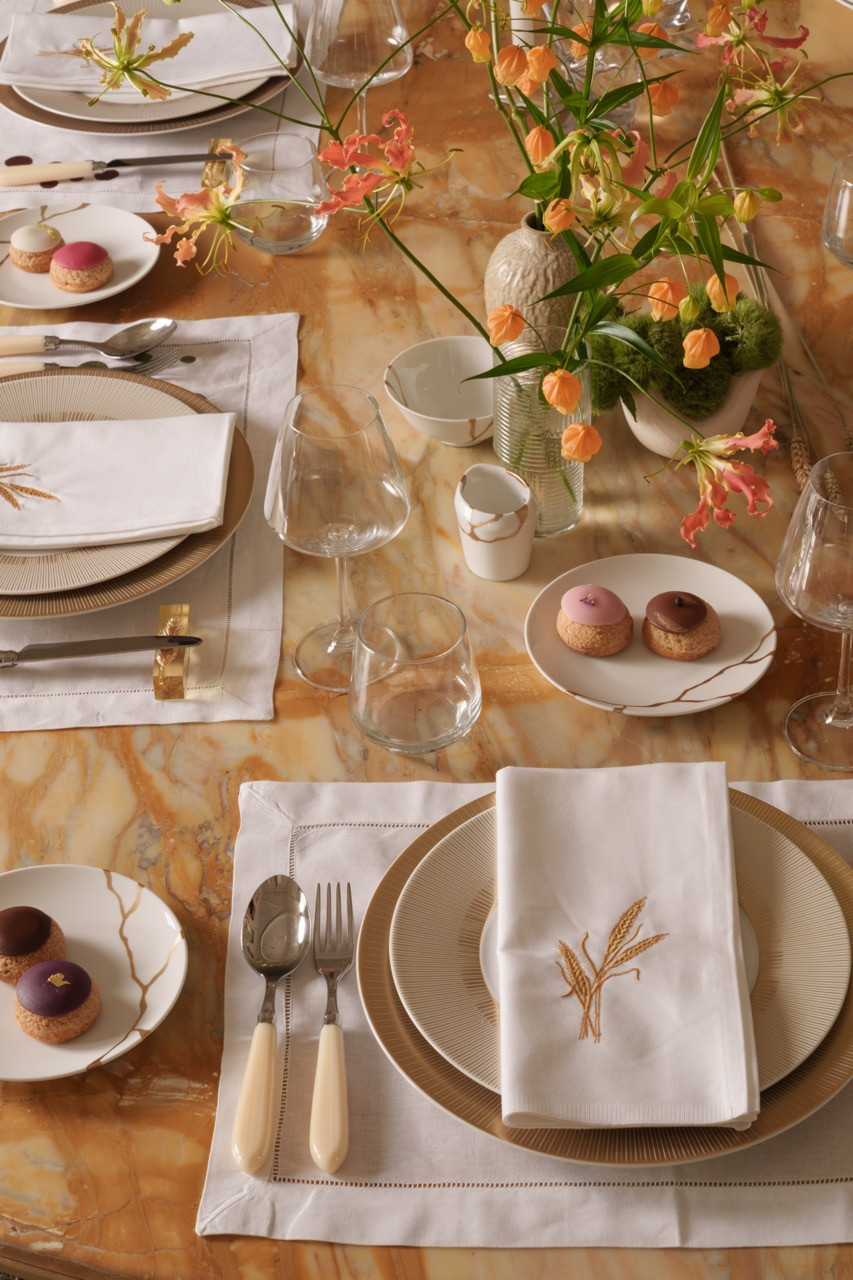 The dining table features custom linens embroidered with wheat motifs that complement the brass lighting fixtures throughout the apartment, set on a travertine surface that Ashby describes as creating a cohesive flow throughout the space